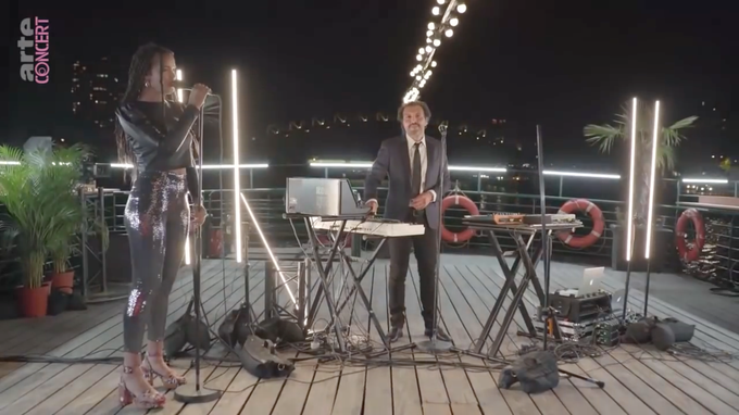 Still frame from the video, a scene on the deck of a boat with multiple keyboards with Kid Francescoli standing between them, and singer Andréa Durand in the front